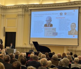 Prelekcja „Działalność Biura Odbudowy Stolicy 1945–1951” kustosza Macieja Aleksiejuka. Na kolorowej fotografii widać przemawiającego do grupy ludzi mężczyznę, w tle prezentacja, fortepian i sprzęt komputerowy.
