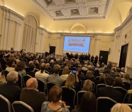 Występ Chóru Kameralnego Tibi Domine z utworem „Budujemy nowy dom”. Na kolorowej fotografii widać grupę muzyków występujących dla licznie zgromadzonej publiczności, obok sprzęt komputerowy i fortepian, na którym gra mężczyzna.