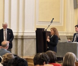 Przemawia Pani Magdalena Wiercińska, dyrektor Archiwum Państwowego w Warszawie. Na kolorowej fotografii widać przemawiającą do grupy ludzi kobietę, obok niej stoi starszy mężczyzna, a za stołem siedzi młody mężczyzna z laptopem.