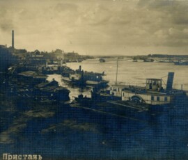 Pocztówka ukazująca przystań w Kijowie, 1917 r., Dar Macieja Aleksiejuka, Archiwum Państwowe w Warszawie, sygn. 72/3882/0/24/1603 Czarno-białe zdjęcie ukazujące brzeg z małymi domkami i przybijające do brzegu statki, w tle woda z pływającymi po niej statkami, w oddali także dwa kominy.