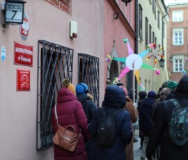 Staromiejskie Kolędowanie w Archiwum Państwowym w Warszawie
Fot. A. Smyk Na kolorowym zdjęciu widać grupę ludzi w kurtkach z torbami i plecakami, jedna z osób trzyma papierową gwiazdę na patyku, obok widać budynek z tablicą Archiwum Państwowe w Warszawie i godłem Orła Białego oraz tabliczkami z tekstami w języku polskim.
