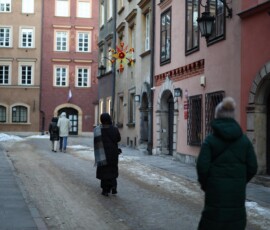 Staromiejskie Kolędowanie w Archiwum Państwowym w Warszawie
Fot. A. Smyk Na kolorowym zdjęciu widać osobę w kurtce z papierową gwiazdą na patyku, idącą w stronę budynku z czerwonymi tablicami z tekstem w języku polskim i godłem Orła Białego, w tle widać inne budynki.