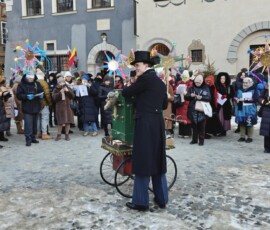 Staromiejskie Kolędowanie z Kataryniarzem Janem na Rynku Starego Miasta
Fot. A. Smyk Na kolorowym zdjęciu widać młodego mężczyznę grającego na katarynce, a wokół niego grupę ludzi w kurtkach i czapkach, przyglądających się jego występowi, w tle widać budynki.
